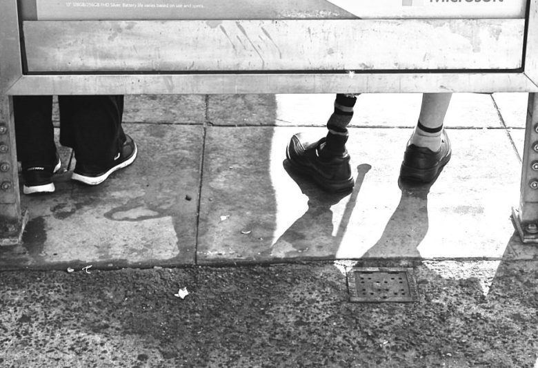 Two people sit at a bus stop; only their legs are visible. One person has a prosthetic leg. Sunlight casts shadows on the pavement.