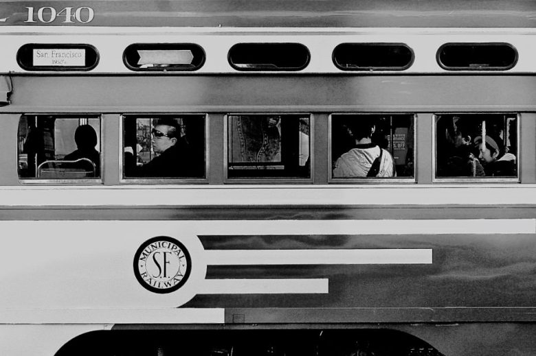 Black and white photo of passengers sitting inside a San Francisco Municipal Railway streetcar, viewed through its windows.