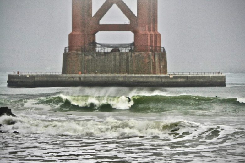 A surfer rides a wave near large red bridge supports on a misty day, with choppy water and a concrete pier in the background.
