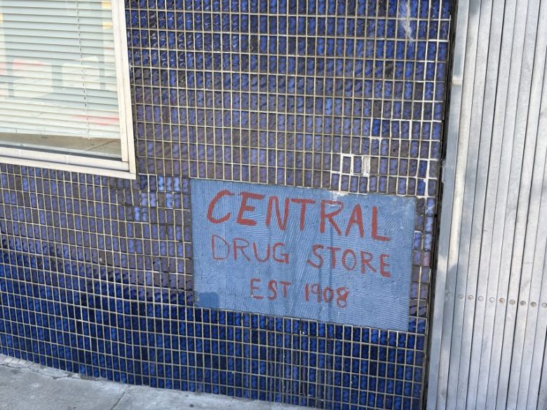 Blue-tiled wall with a hand-painted sign reading “CENTRAL DRUG STORE EST 1908” in red and white letters, next to a window with closed blinds.