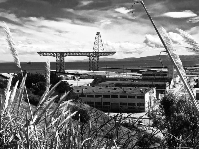 Black-and-white photo of an industrial waterfront site with large steel structure, warehouses, and tall grass in the foreground under a partly cloudy sky.