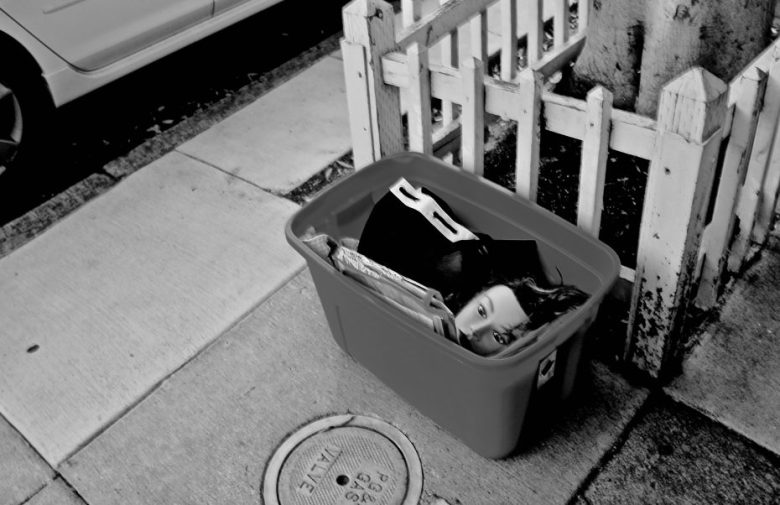 A plastic storage bin sits on a sidewalk near a white fence, containing newspapers and a doll’s head, partially concealed.