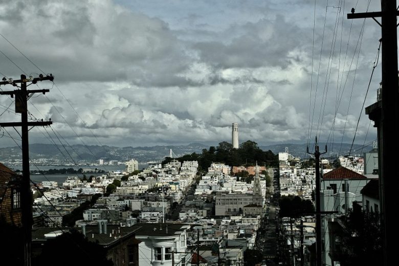 A cityscape view of San Francisco with the Coit Tower on a hill, dense buildings, power lines, and cloudy skies.