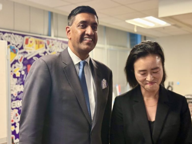 Two people in business attire stand indoors, one smiling at the camera, the other looking downward with a slight smile. A colorful mural is visible in the background.