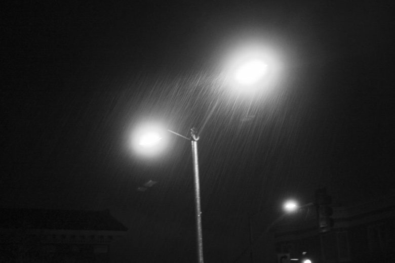 A streetlamp glows at night, illuminating falling rain. Two building rooftops and a traffic light are faintly visible in the dark background.