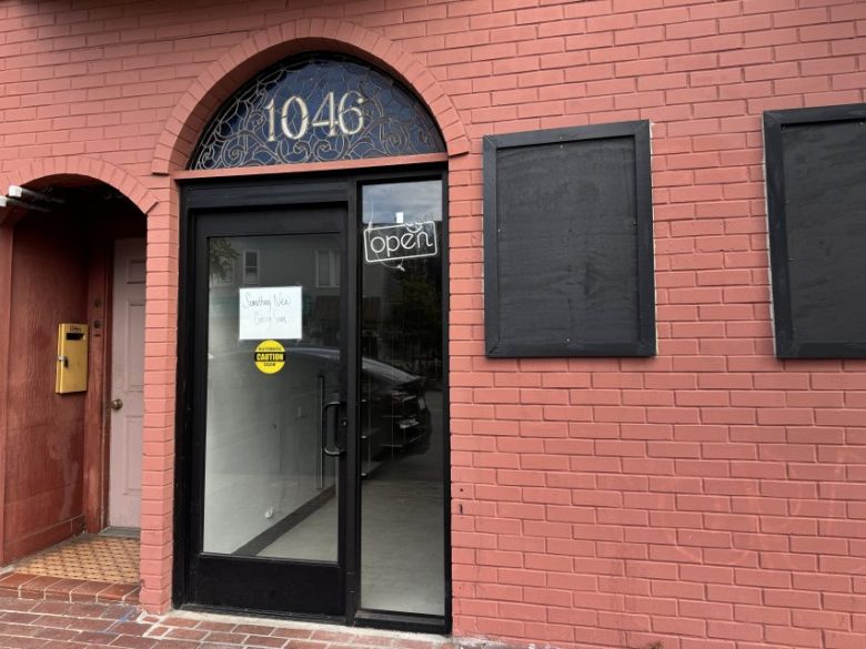 A red brick storefront at 1046 with a glass door displaying an “open” sign and a handwritten note that says “Southern Vegan coming soon.” Two window spaces are covered.