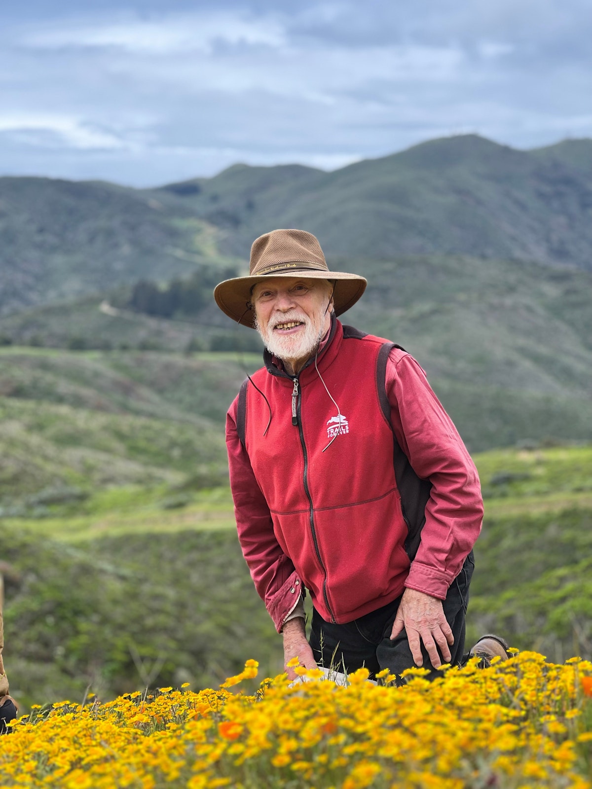 Jake Sigg kneels amid a swath of California goldfields (Lasthenia californica) during a tour of the Crystal Springs Reservoir led by the Public Utilities Commission. In the background are Montara Mountain and coastal grassland prairies. The photo was taken May 5, 2023. 