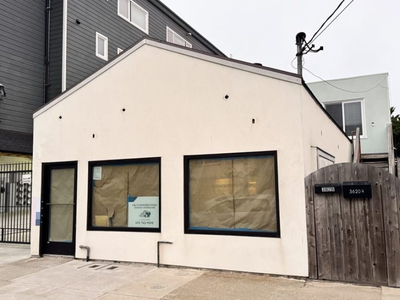 A small, white, single-story building with covered windows and a wooden gate on the right marked “3602B.” A construction sign is posted in one window.