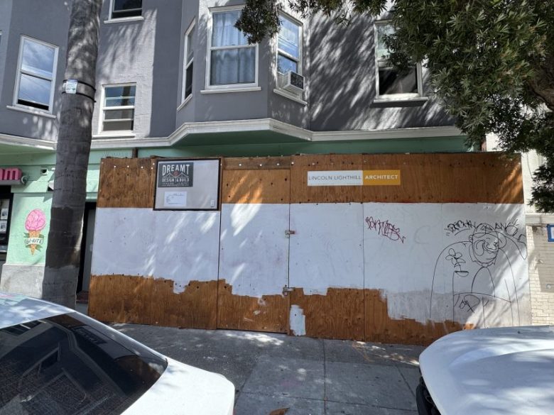 Plywood construction barrier covers a building facade with graffiti, an architect sign, and a framed notice. Trees and parked cars are visible at the edges.