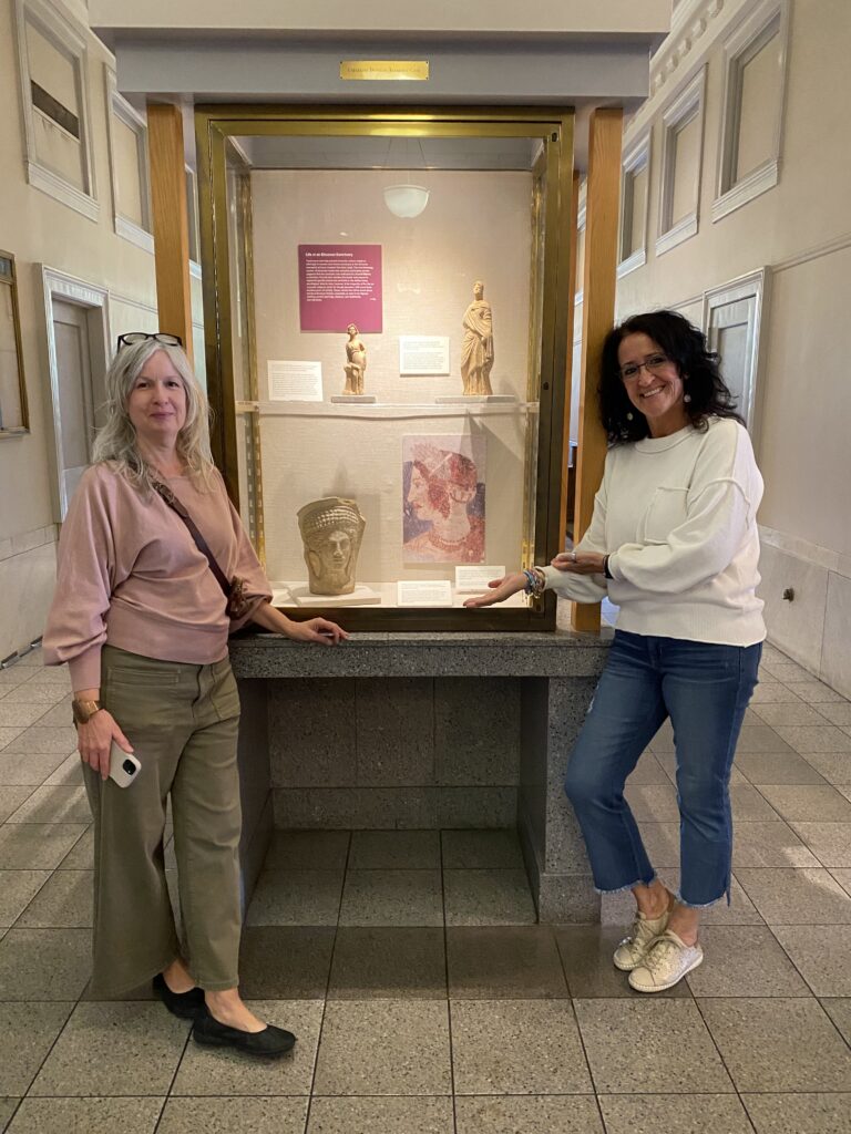 Two people stand next to a glass display case that contains ancient pottery. The cases are in a lobby, and both people are looking toward the camera.