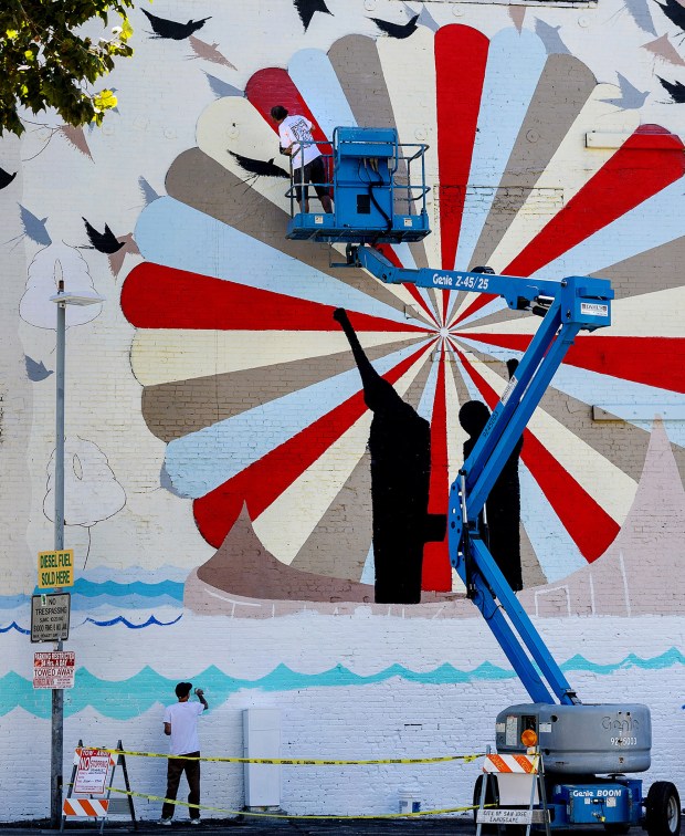 The "Homage" mural celebrating Olympic sprinters John Carlos and Tommie Smith next to the Chevron Station on Santa Clara and Fourth Streets in San Jose, Calif., is touched up in August of 2024, by original artist Paul Urich (lower left), with the help of Brian Eder (in the cherry picker), Steven Reece and Cherri Lakey. (Karl Mondon/ Bay Area News Group)