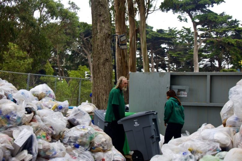 Two people in green jackets stand among piles of bagged trash and recycling bins near a dumpster in an outdoor, wooded area.