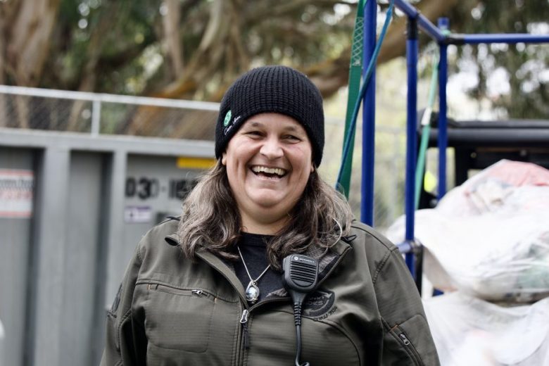 A person wearing a black beanie and green jacket with a radio clipped on, smiling outdoors near a fence and bags of gathered items.