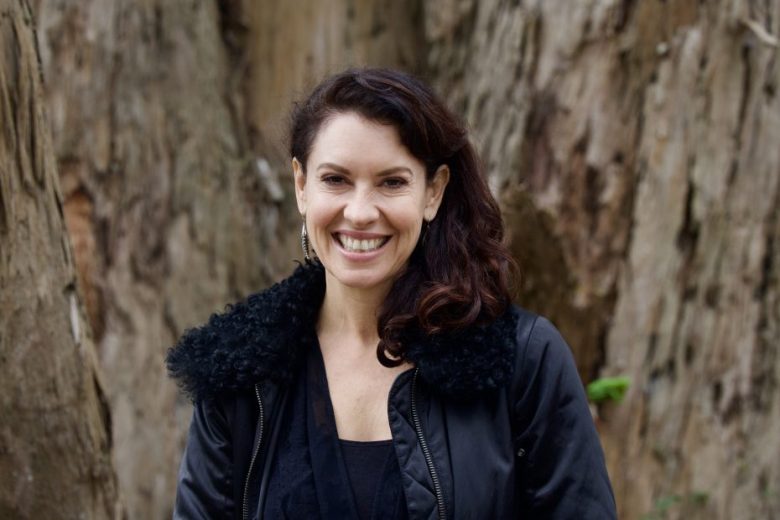 Woman with dark hair wearing a black jacket stands outdoors in front of a large tree trunk, smiling directly at the camera.
