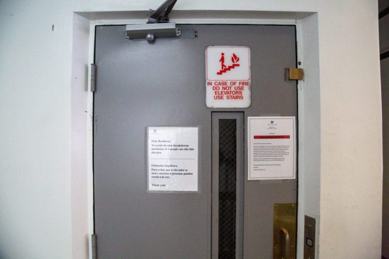 Gray elevator door with fire safety sign above, white notice and paper on the door, and an elevator button panel on the right side of the door frame.