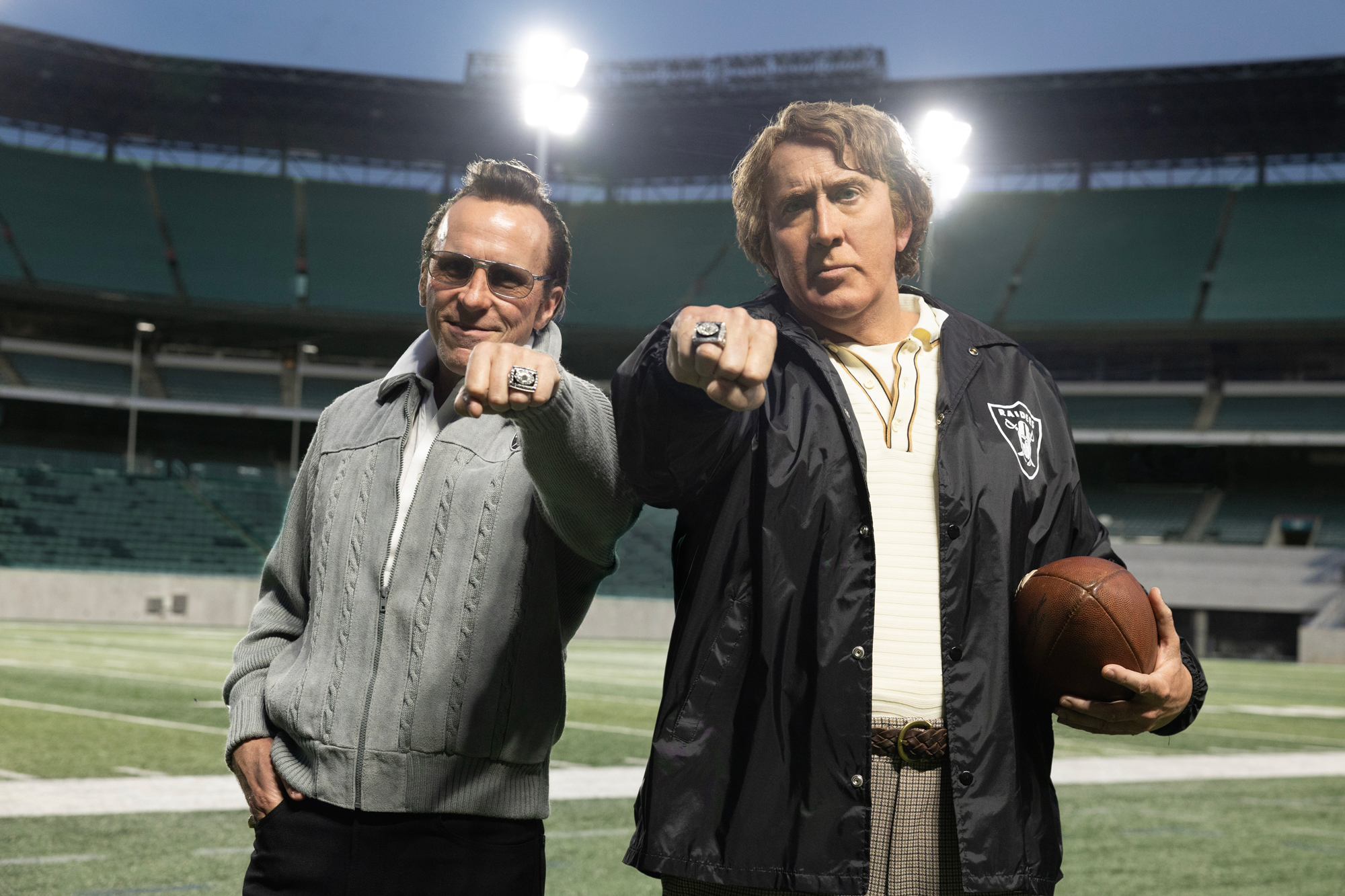 two men on football field punch fists toward camera with championship rings on fingers, one holds a football