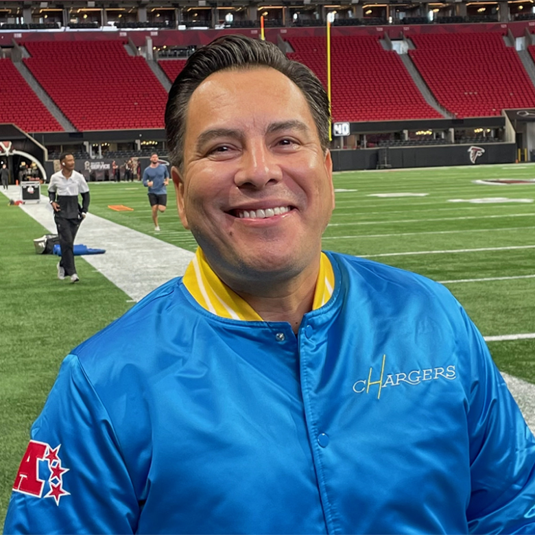 Smiling man in a bright blue Chargers jacket poses on an NFL football field, with green turf and red stadium seats behind him.