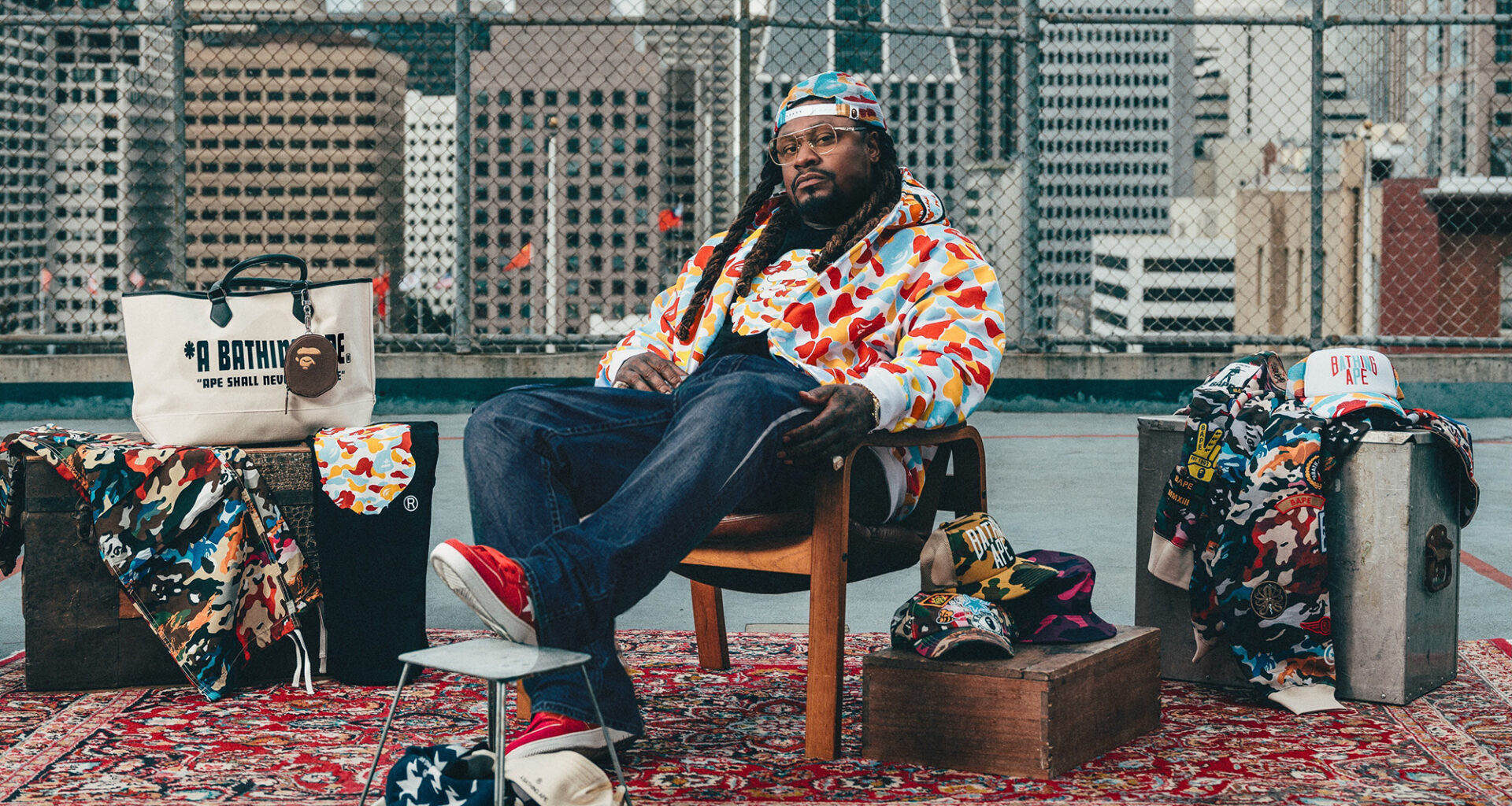 A Black man in glasses, backwards cap, jeans and colorful patterned jacket sits in a chair on an oriental rug with a backdrop of chain-link fencing and the San Francisco skyline