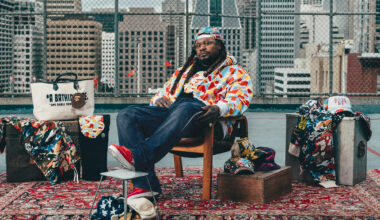 A Black man in glasses, backwards cap, jeans and colorful patterned jacket sits in a chair on an oriental rug with a backdrop of chain-link fencing and the San Francisco skyline