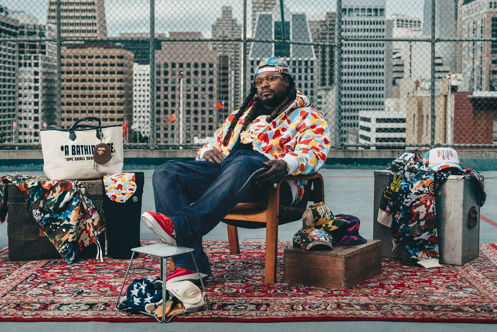 A Black man in glasses, backwards cap, jeans and colorful patterned jacket sits in a chair on an oriental rug with a backdrop of chain-link fencing and the San Francisco skyline
