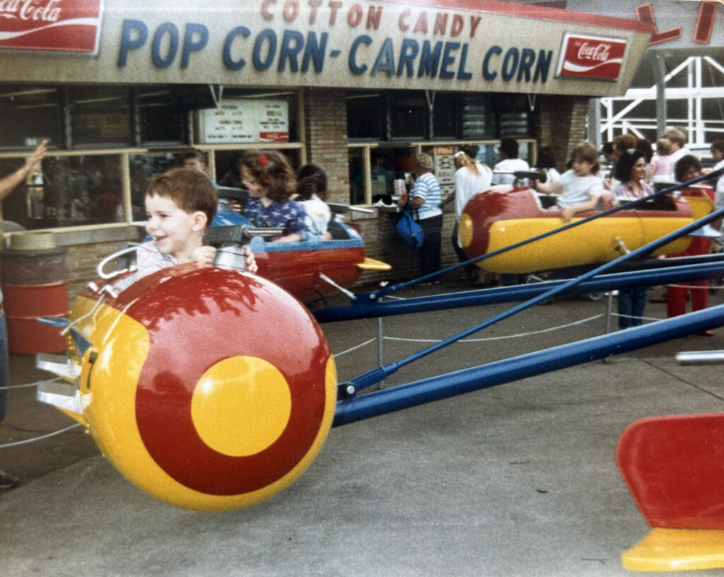 Vintage photograph of a smiling young boy riding a red-and-yellow airplane carnival ride in front of a concession stand.