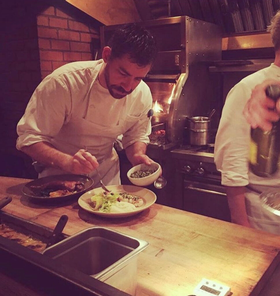 Michael Battisto, dressed in a chef's coat, intently plates food at a wooden kitchen pass in front of a wood-fired oven.