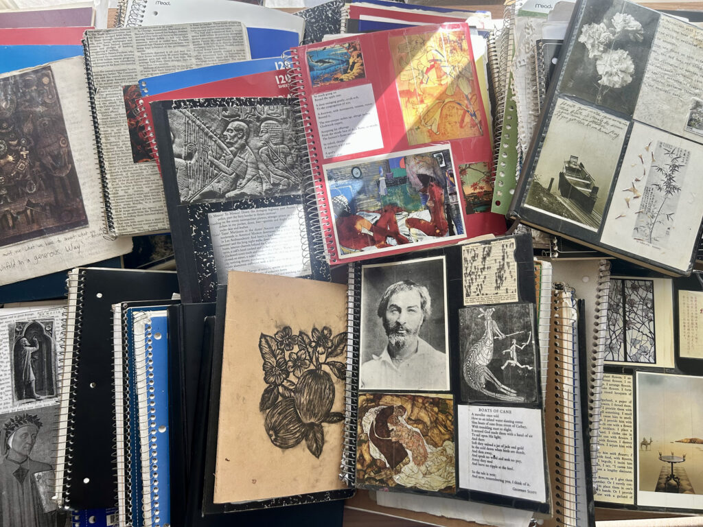 A top-down collection of stacked wire-bound notebooks and journals, decorated with pasted photos, artwork, newspaper clippings, and handwritten text.
