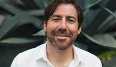 Close-up portrait of a smiling Michael Battisto wearing a white collared shirt against a background of dark green agave leaves.