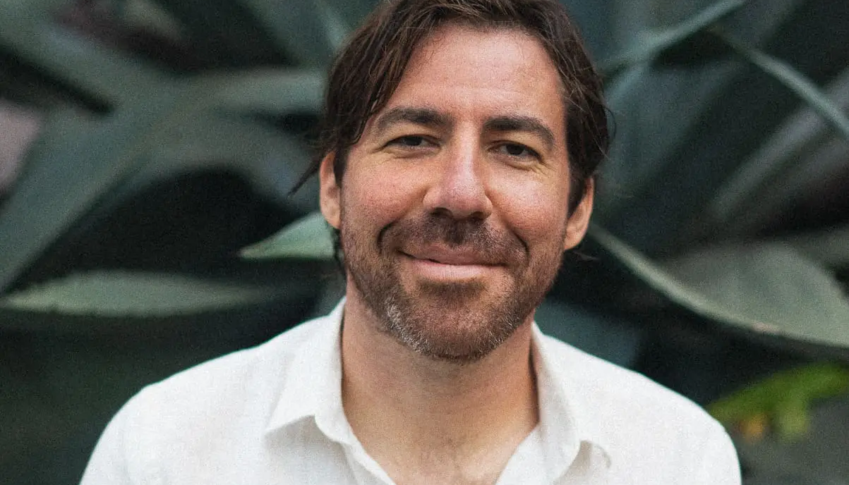 Close-up portrait of a smiling Michael Battisto wearing a white collared shirt against a background of dark green agave leaves.