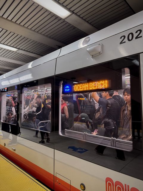 A light rail train labeled “Ocean Beach” is at a station. Passengers are seated and standing inside the train, while others wait on the platform.