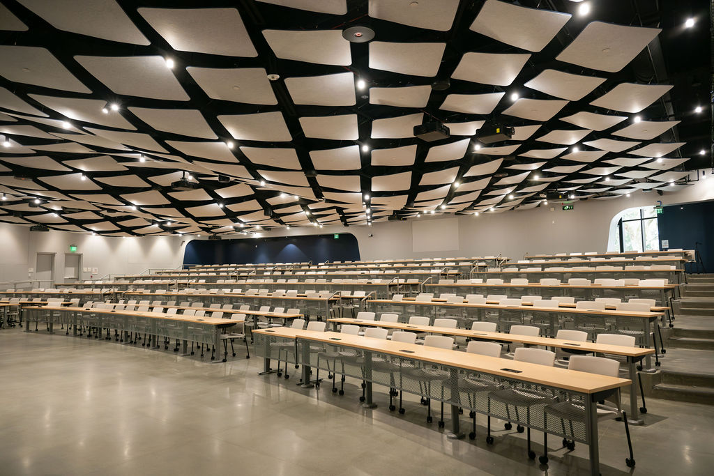 A view of the new auditorium inside the Undergraduate Academic Building, looking toward the back from the front corner. The room is empty, with hundreds of chairs pushed up to the desks. Wavy, angular ceiling panels give the room a unique, modern appearance.