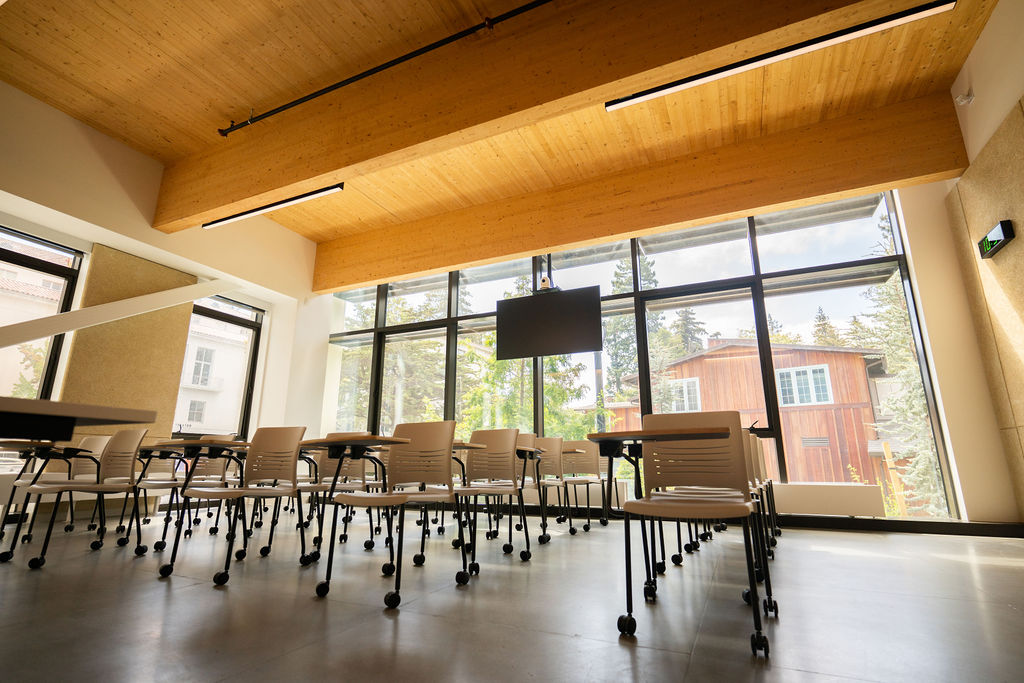 A brightly lit classroom, with floor-to-ceiling windows on two sides, giving the room ample bright, natural light. Wood beams and high ceilings give the room an open feeling, while the chairs are lined in rows.