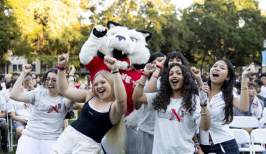 Students cheer with arms raised alongside Northeastern's husky mascot at an outdoor welcome event, with a large crowd seated behind them.