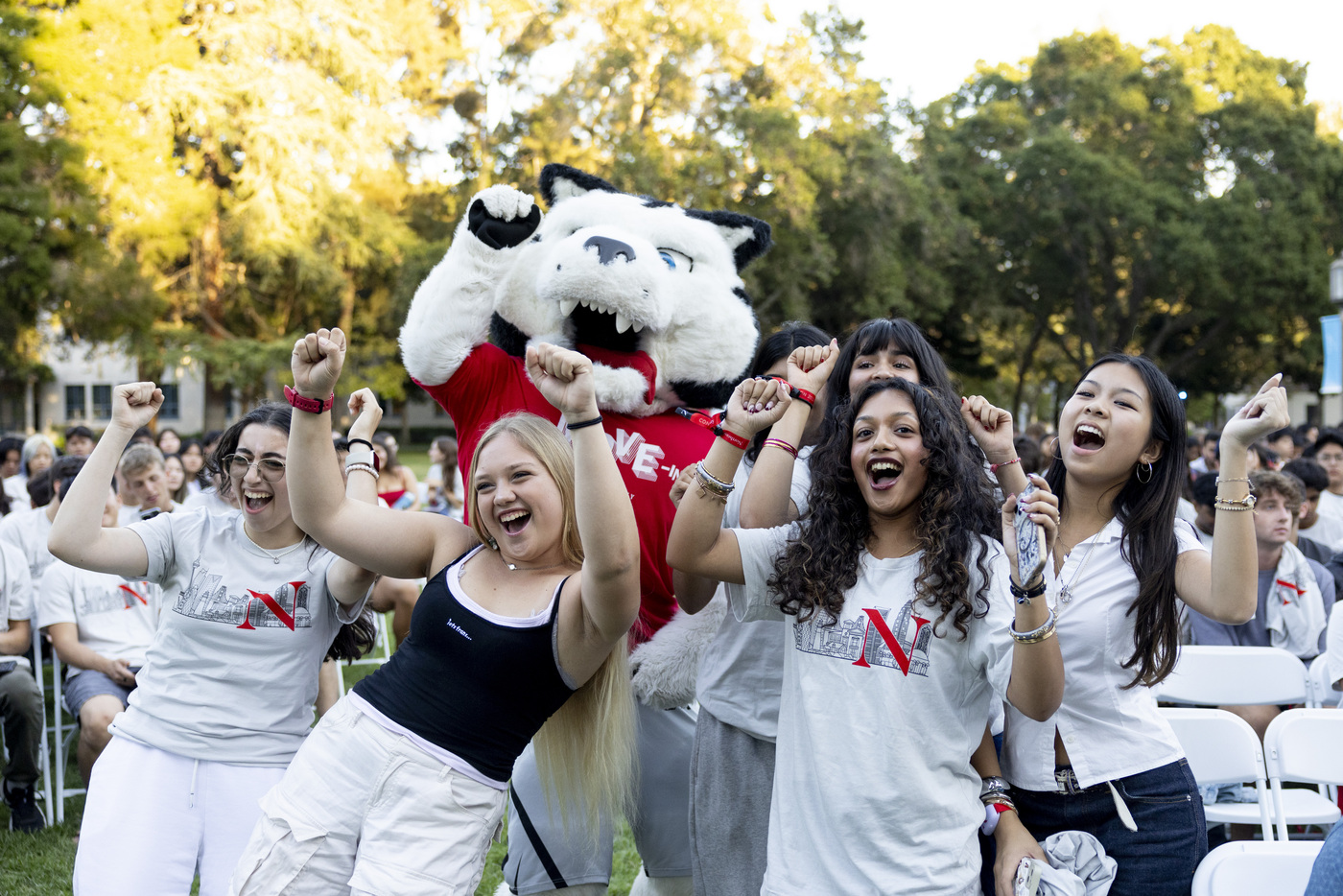 Students cheer with arms raised alongside Northeastern's husky mascot at an outdoor welcome event, with a large crowd seated behind them.