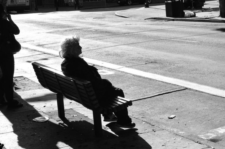 An elderly person sits alone on a bench by a quiet, sunlit sidewalk, casting a long shadow across the pavement.