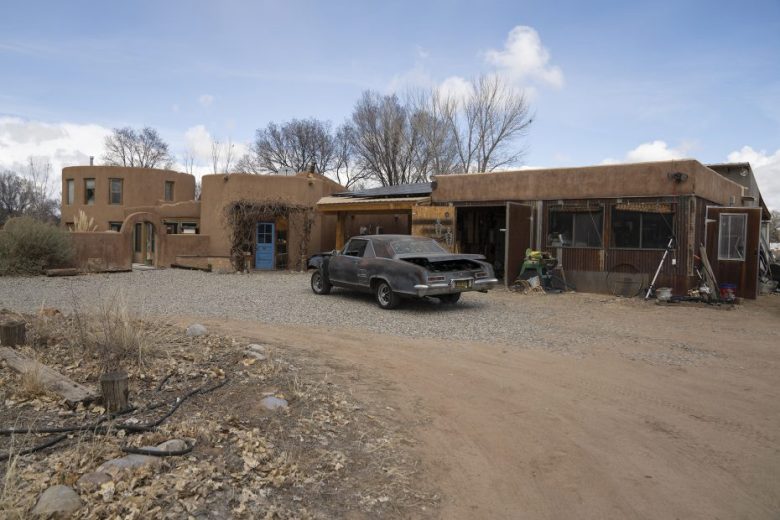 A rustic adobe-style house with multiple sections, a gravel driveway, and an old black car parked in front, surrounded by leafless trees and dry ground.