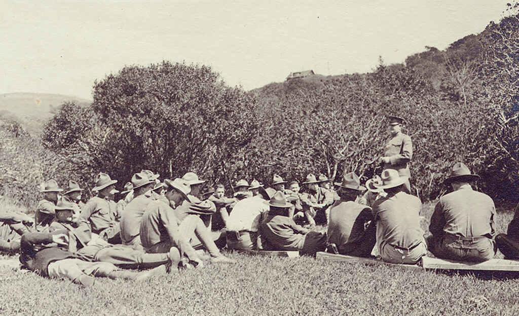 In a sepia-toned photograph from 1917, an Army officer stands among a group of roughly 30 UC Berkeley ROTC cadets who are sitting on the ground and listening.