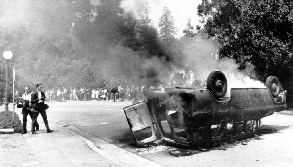 In this black-and-white photo from 1970, a van, upside down in the road, emits flames and smoke as bystanders keep their distance.