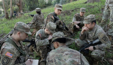 A group of eight ROTC cadets in military garb crouch in the green grass of a wooded hillside as they consult about their training exercise