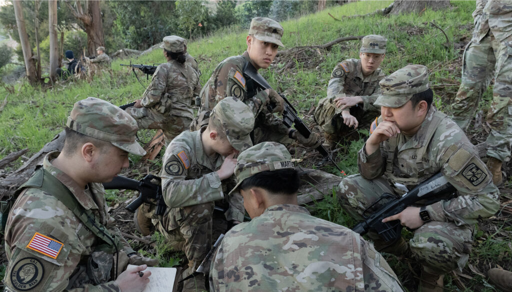 A group of eight ROTC cadets in military garb crouch in the green grass of a wooded hillside as they consult about their training exercise
