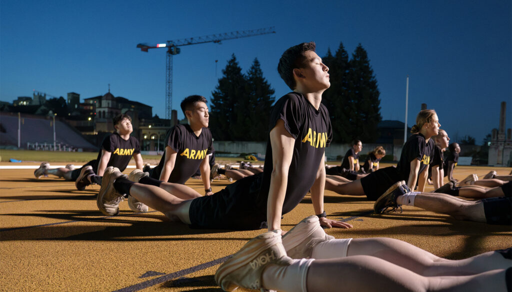 In the mid-morning light, a group of cadets wearing black t-shirts emblazoned with the word "Army" do stretching exercises after a physical training session