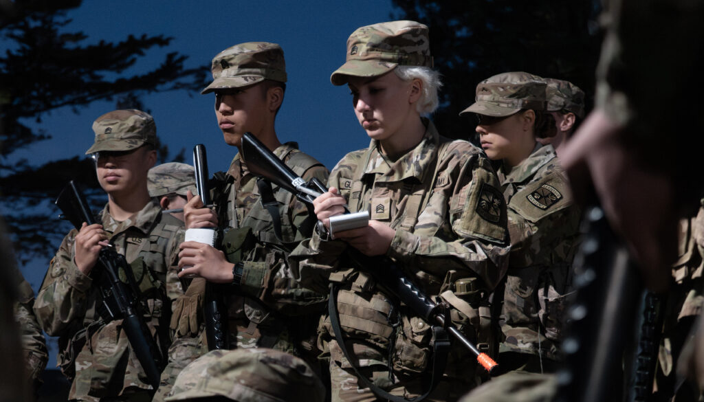 A group of Army ROTC cadets in camouflage military uniforms and hats stand as they listen to a pre-dawn briefing.