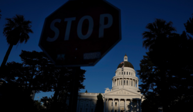 A dimly lit stop sign is in the foreground with the brightly sunlit dome and facade of a classical government building behind it, framed by tall trees.