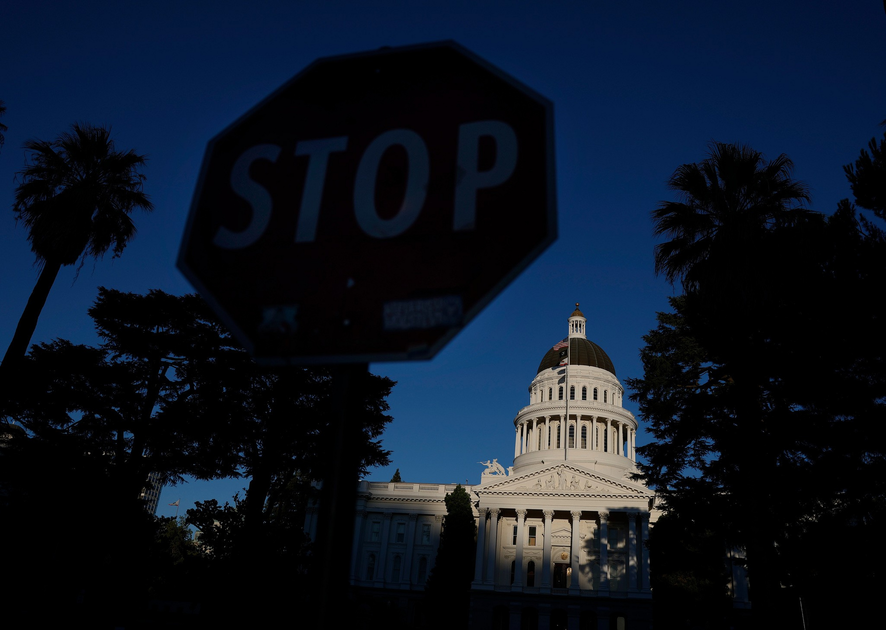 A dimly lit stop sign is in the foreground with the brightly sunlit dome and facade of a classical government building behind it, framed by tall trees.