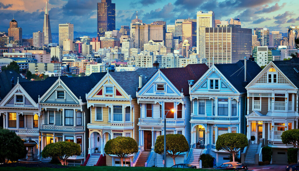 The famous "painted ladies," a row of Victorian-era houses in San Francisco with the city's downtown area in the background, under a dramatic sky.