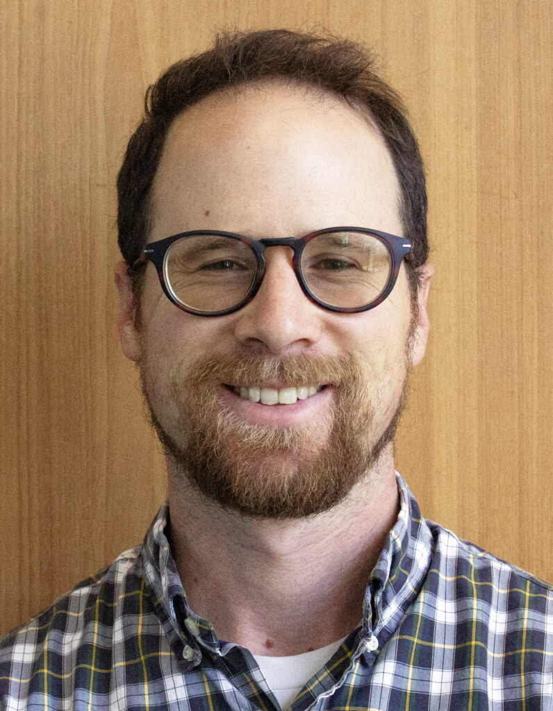 headshot of Sam Trachtman, smiling, against a wood-panelled backdrop