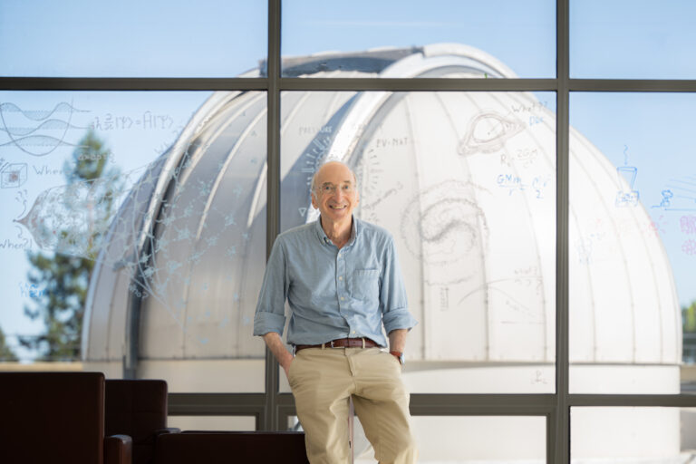 Saul Perlmutter leans in front of a window with equations and drawings, with a telescope in the background.