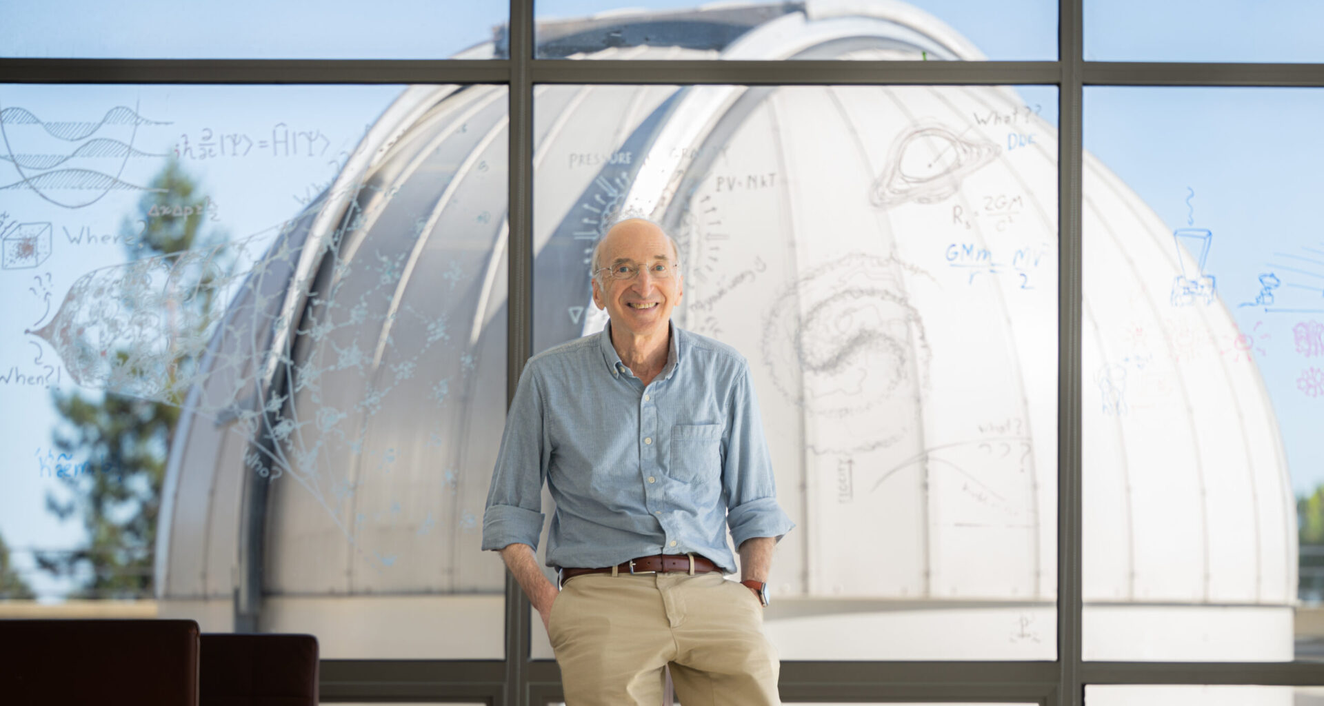 Saul Perlmutter leans in front of a window with equations and drawings, with a telescope in the background.