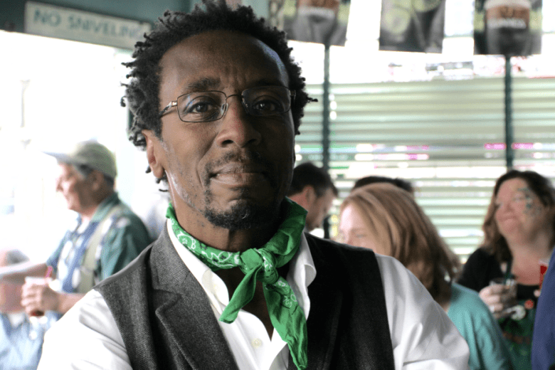 Man with glasses and a green bandana around his neck stands indoors with a group of people in the background.