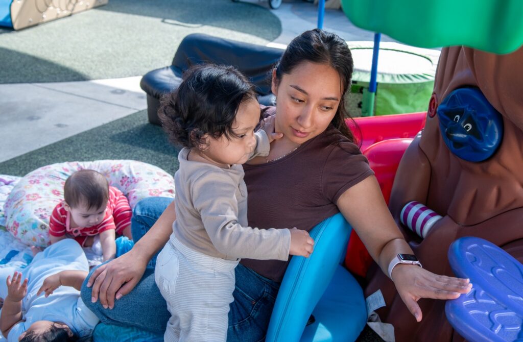A Latina educator sits outdoors on a low blue floor chair, supporting a toddler wearing a beige long-sleeved shirt who stands in her lap and leans on her for balance. The adult reaches out her hand toward a brown treehouse-style plastic play structure. Next to them on a patterned blanket, one infant and one child play on the ground: the baby in a red striped shirt rests against a floral nursing pillow, while another in a light blue outfit lies on their back looking upward.